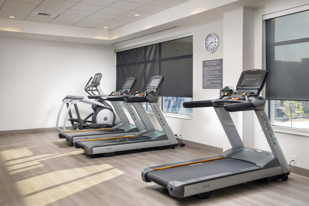 Two treadmills and an elliptical machine lined up in a fitness room with a wall-mounted clock and posted exercise rules.