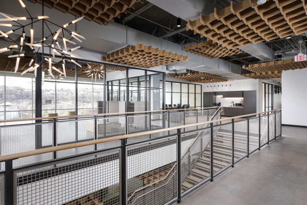 A modern office interior featuring a mezzanine walkway with metal railings overlooking a staircase, set beneath a waffle-patterned wooden ceiling.