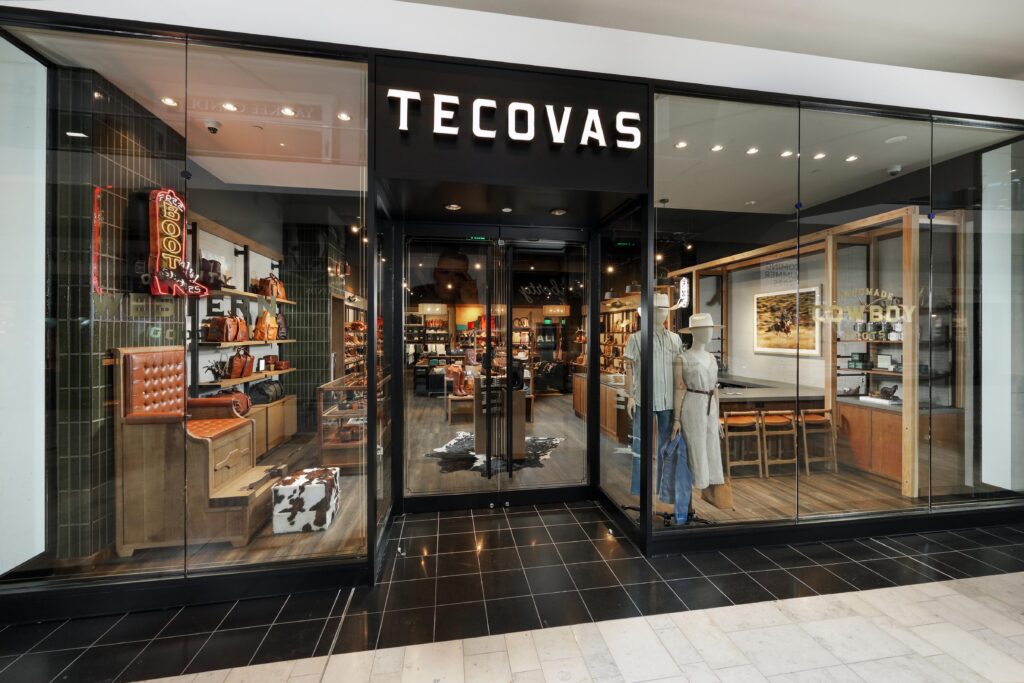 Storefront of a Tecovas retail shop with mannequins and leather goods visible through the glass windows.