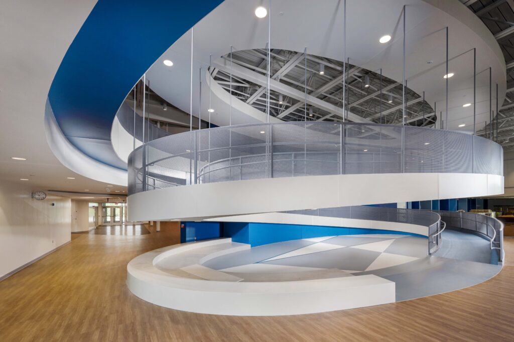 A wide, white spiral ramp with mesh railings encircles a circular seating area in a large, open room with wood-look flooring and blue accent walls.