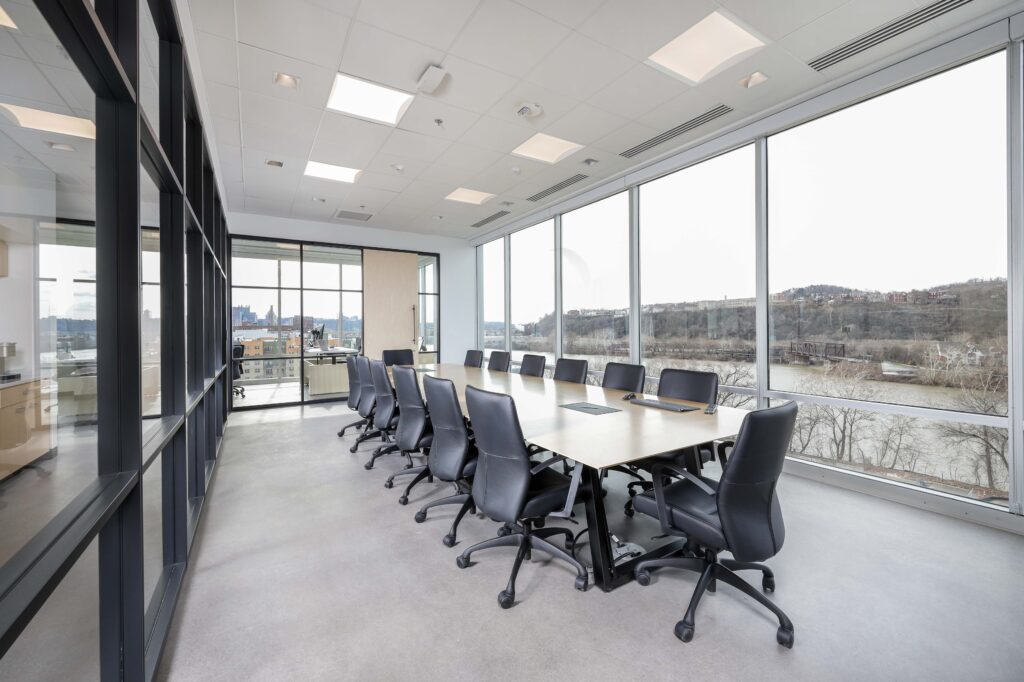 Modern conference room with a long table and black rolling chairs, featuring large windows overlooking a river and city skyline.