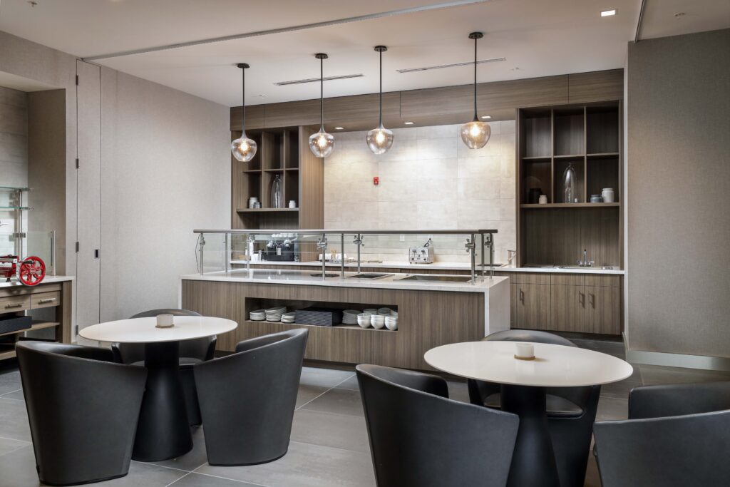 A hotel or office dining area featuring two round tables with black chairs in the foreground and a service counter with shelving and pendant lights in the background.