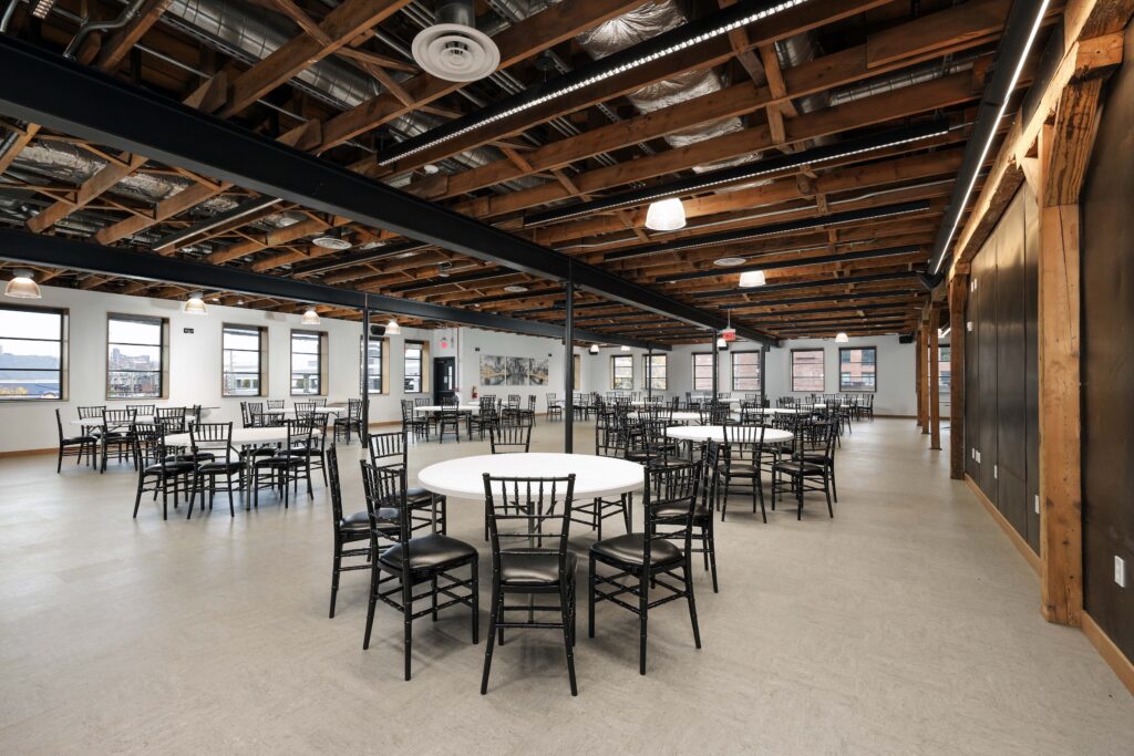 Large event space with exposed wooden beams, white walls, and multiple round tables set with black chairs.