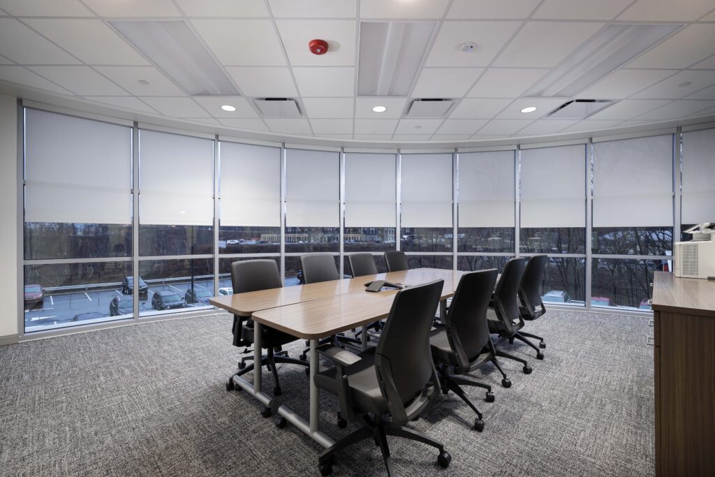 A conference room featuring a long wooden table surrounded by black office chairs, set against a curved wall of windows with lowered shades.