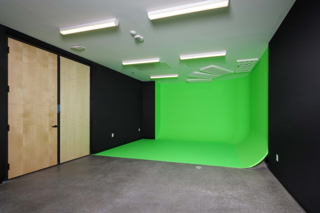 A professional studio space featuring a large green screen cyclorama wall, a concrete floor, and light wood double doors.
