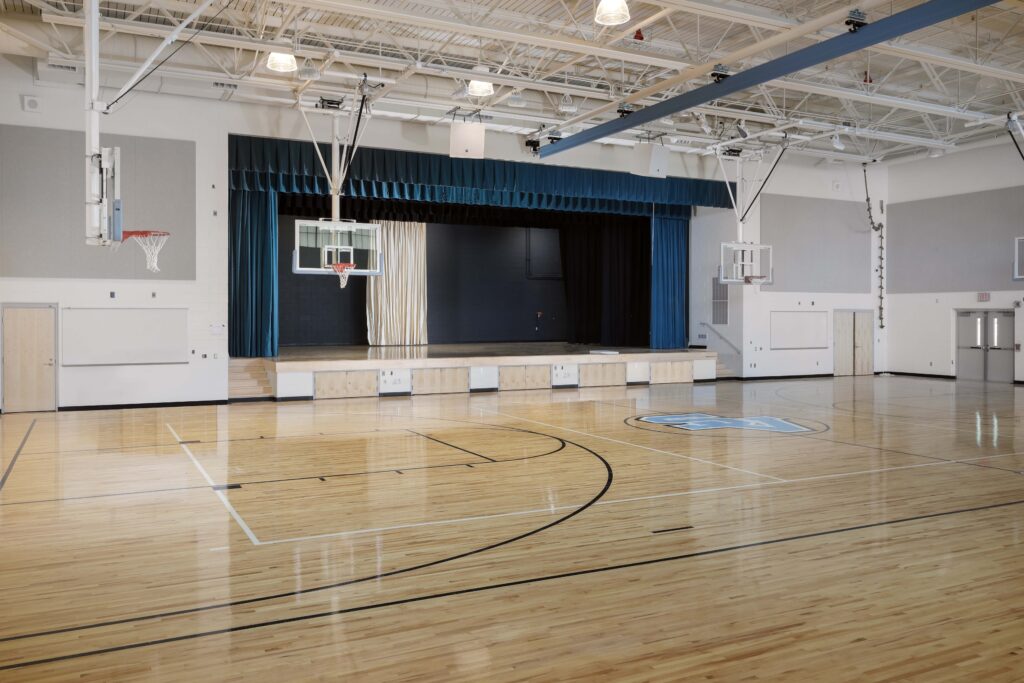 A gymnasium with a hardwood floor and basketball hoops, featuring a raised stage with blue curtains at the far end.
