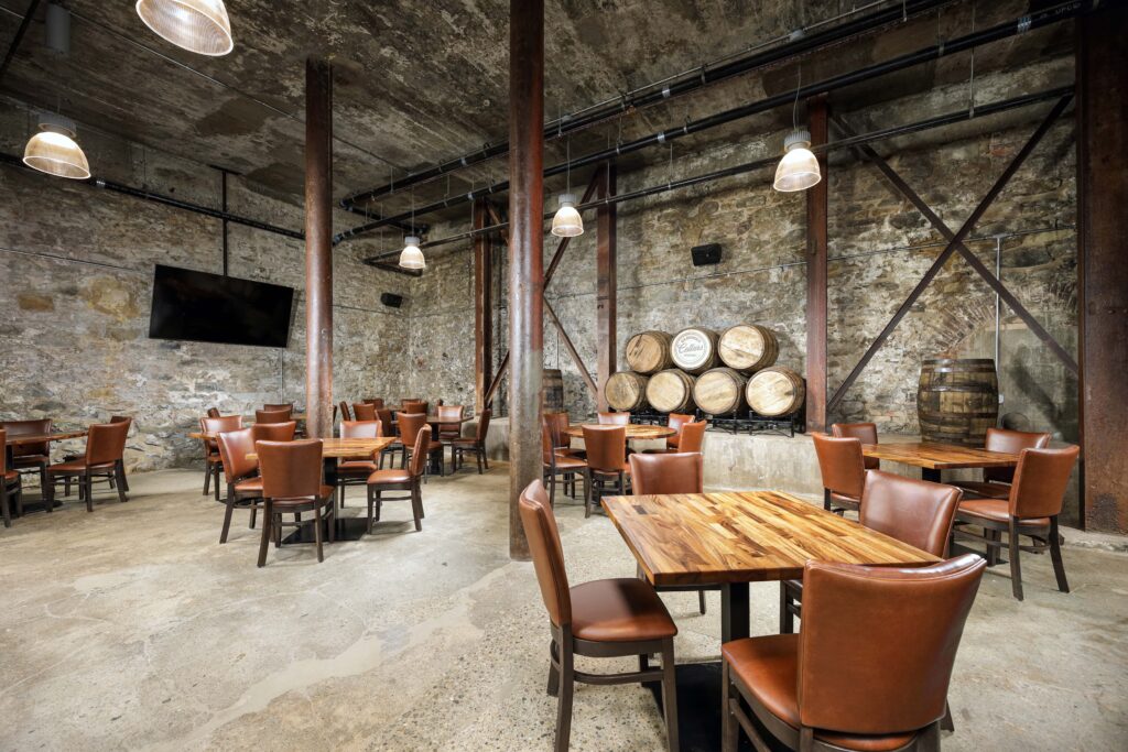 Dining room in a stone-walled cellar with wooden tables, brown chairs, and stacks of barrels.