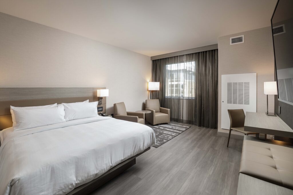 A hotel room with a king bed, two armchairs, a desk with a chair, and floor-to-ceiling windows with curtains.