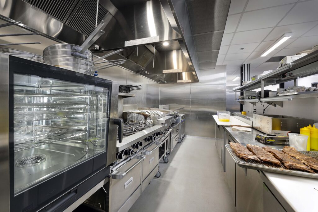 A professional stainless steel kitchen with an oven, stovetop, and a rack of ribs on a prep table.