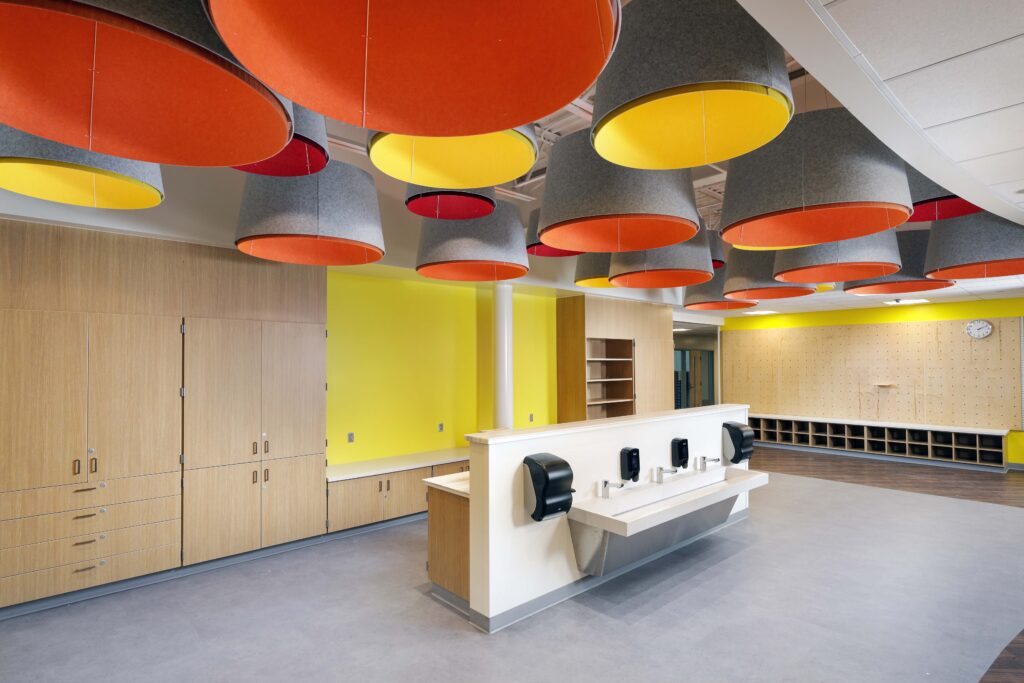 Large, colorful pendant lights hang over a school-style wash station with soap dispensers and a sink, set against yellow walls and wooden cabinetry.