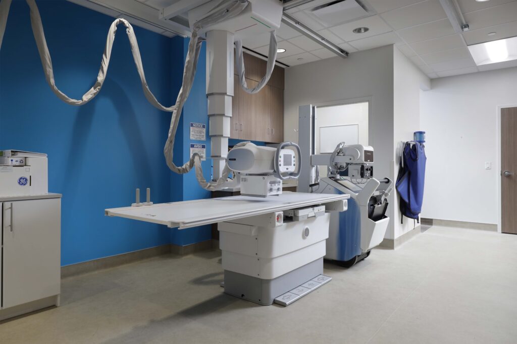 An X-ray imaging room featuring a central examination table with an overhead tube, a mobile X-ray unit, and a blue wall.