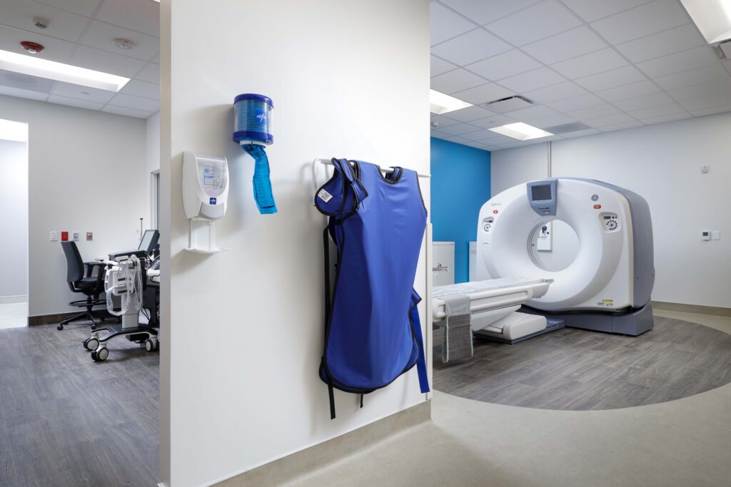 A lead apron hangs on a wall in a medical imaging room containing a CT scanner.