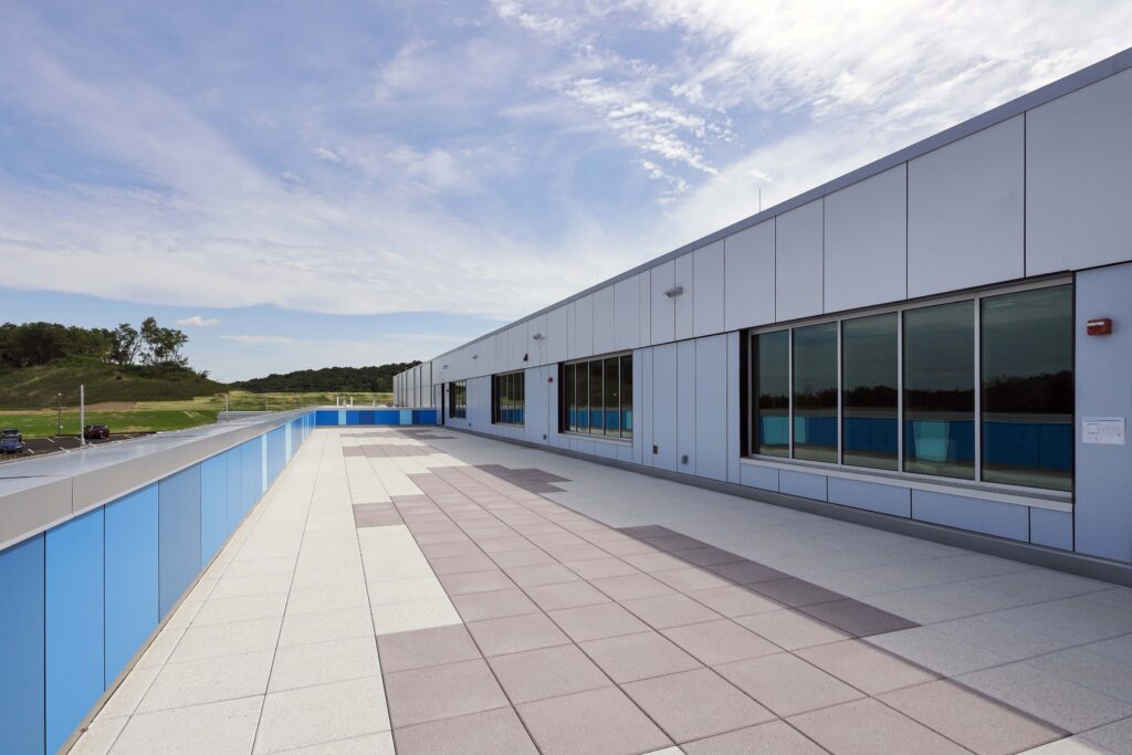 A flat, paved rooftop terrace with large windows along a light-colored metal building, overlooking a grassy hill.