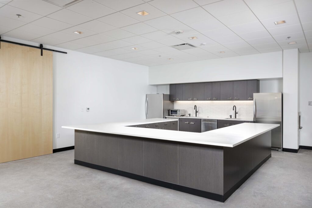 A modern office breakroom featuring a U-shaped kitchen island, dark grey cabinetry, and two stainless steel refrigerators.
