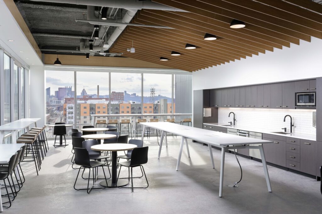 Modern office breakroom with a long white communal table, several small round tables, and a kitchen area, overlooking a city skyline through large windows.