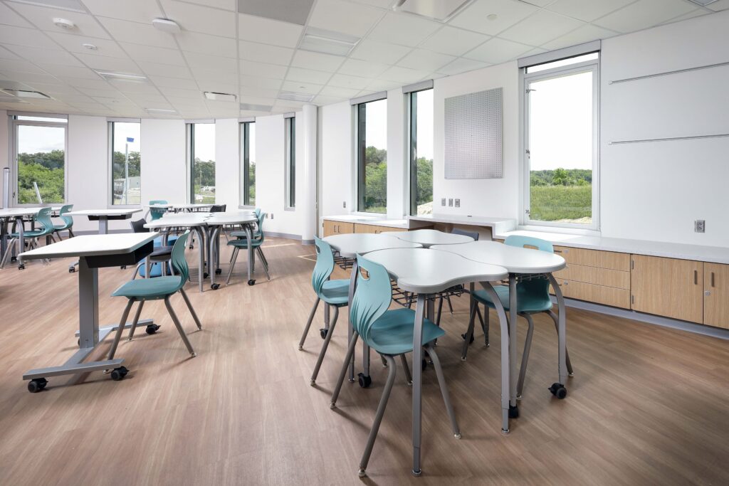A modern classroom with light-colored curved desks and teal chairs arranged on a wood-look floor near floor-to-ceiling windows.