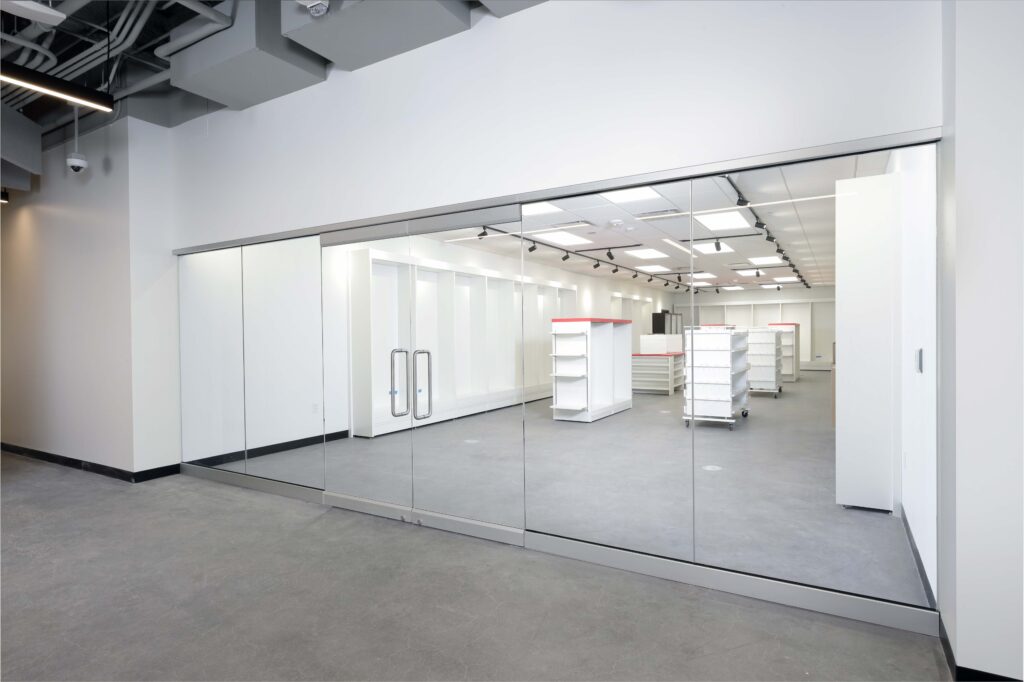 Empty commercial storefront with glass doors, white shelving units, and a concrete floor.