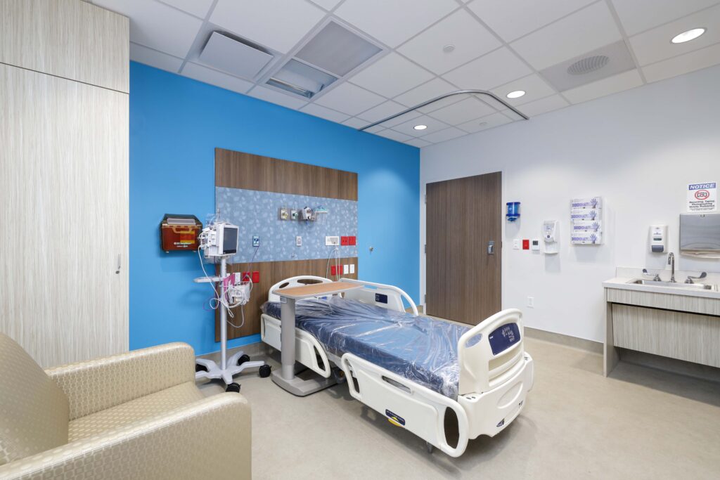 A hospital patient room with an adjustable bed, bedside medical equipment, a chair, and a sink.