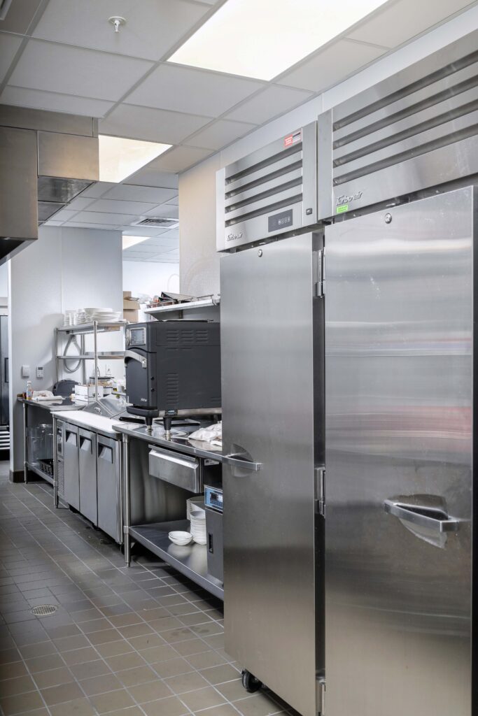 Commercial kitchen interior featuring a large stainless steel refrigerator, food preparation counters, and equipment.