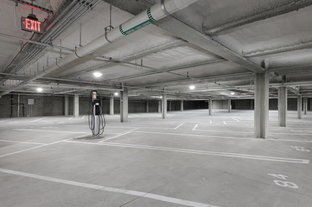 Empty concrete parking garage with a single electric vehicle charging station and an illuminated exit sign.