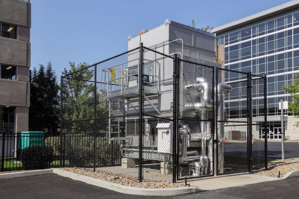 An industrial cooling unit enclosed in a chain-link fence between two office buildings.