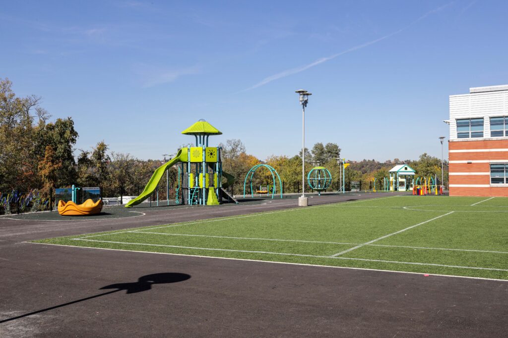 Outdoor playground with slides, climbing structures, and a turf sports field next to a school building.