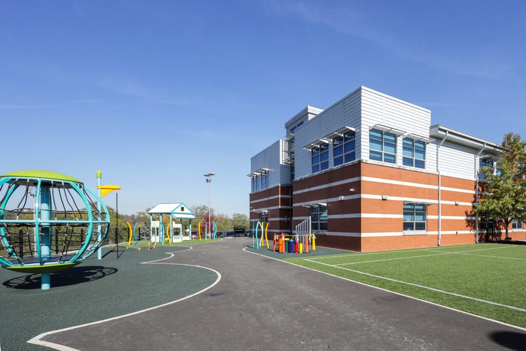 A modern two-story brick and metal school building adjacent to an outdoor playground with a climbing structure and various play equipment.
