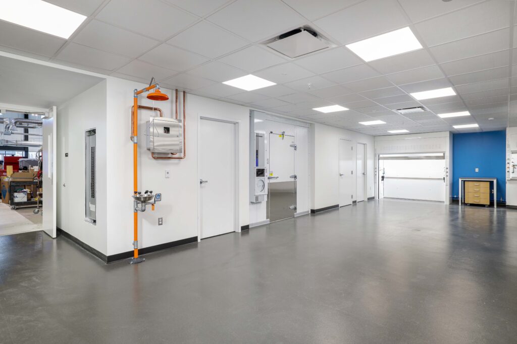 A sterile laboratory hallway featuring an emergency eye wash and shower station, white doors, and a walk-in cooler door.