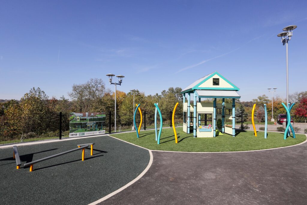 A small playhouse structure on a turf surface at a playground, with a balance beam in the foreground and a sign listing project partners.