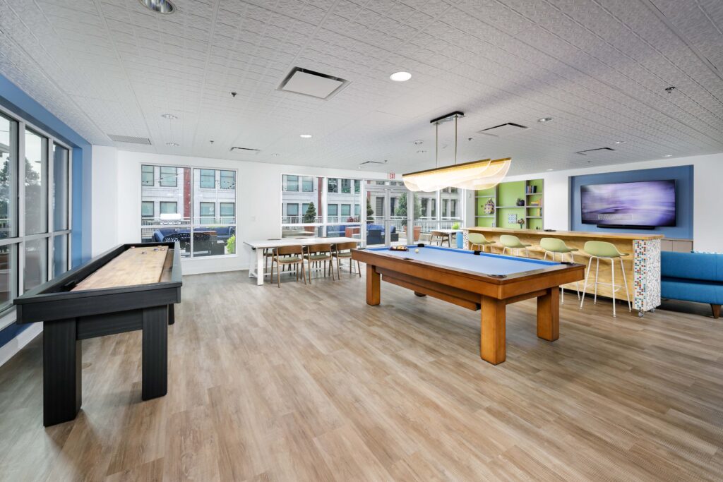 A game room featuring a shuffleboard table, a pool table, a long dining table with chairs, and a bar area with stools in front of a wall-mounted television.