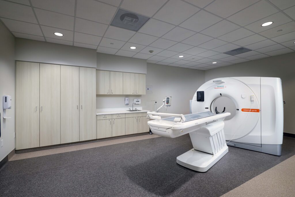 A CT scanner in a clinical exam room with a patient bed, cabinets, and a sink.