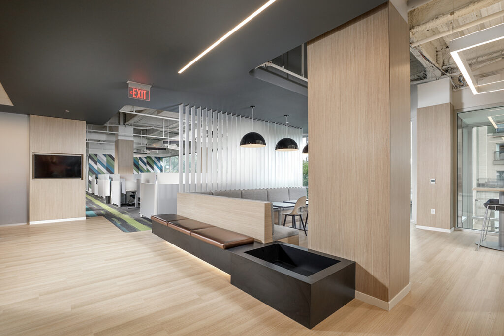 Modern open-plan office space featuring a central wood-paneled pillar, a built-in bench with leather seating, and rows of partitioned cubicles in the background.