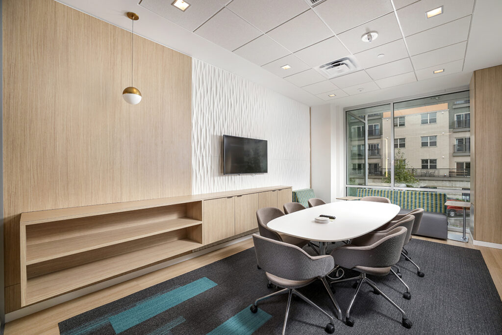 A conference room with an oval table, six grey armchairs, a wall-mounted television, and built-in storage cabinets.