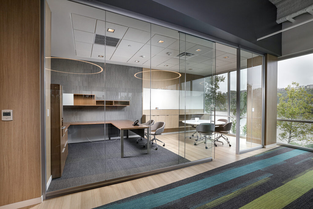 A modern glass-walled office with a wooden L-shaped desk, a round meeting table, and views of trees outside.