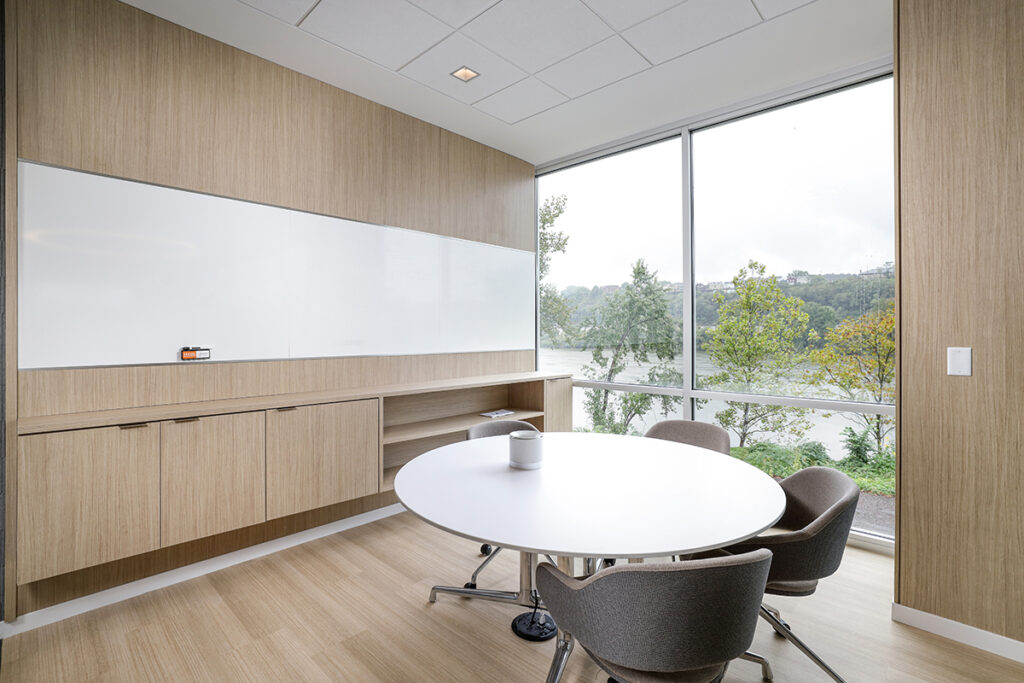 A meeting room with a round table, four chairs, a white board, and a large window overlooking trees and a river.