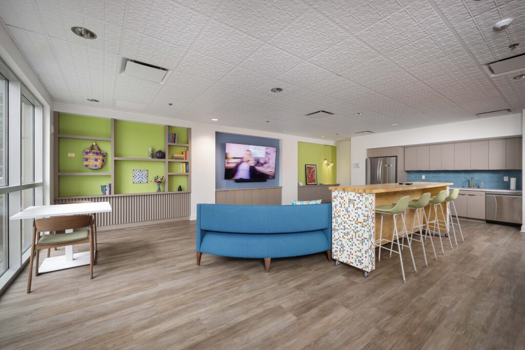 A modern break room featuring a blue curved sofa, a kitchen island with bar stools, and a green wall with built-in shelving.