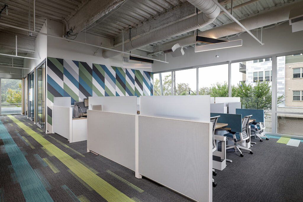 Office interior with rows of cubicles, patterned wall, and floor-to-ceiling windows overlooking a building.