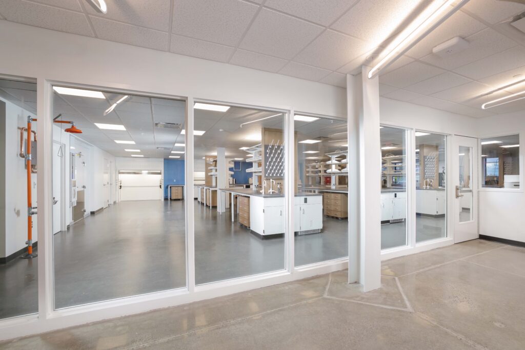 A bright laboratory space viewed through a glass wall, featuring rows of white workbenches, metal cabinets, and an emergency shower station.