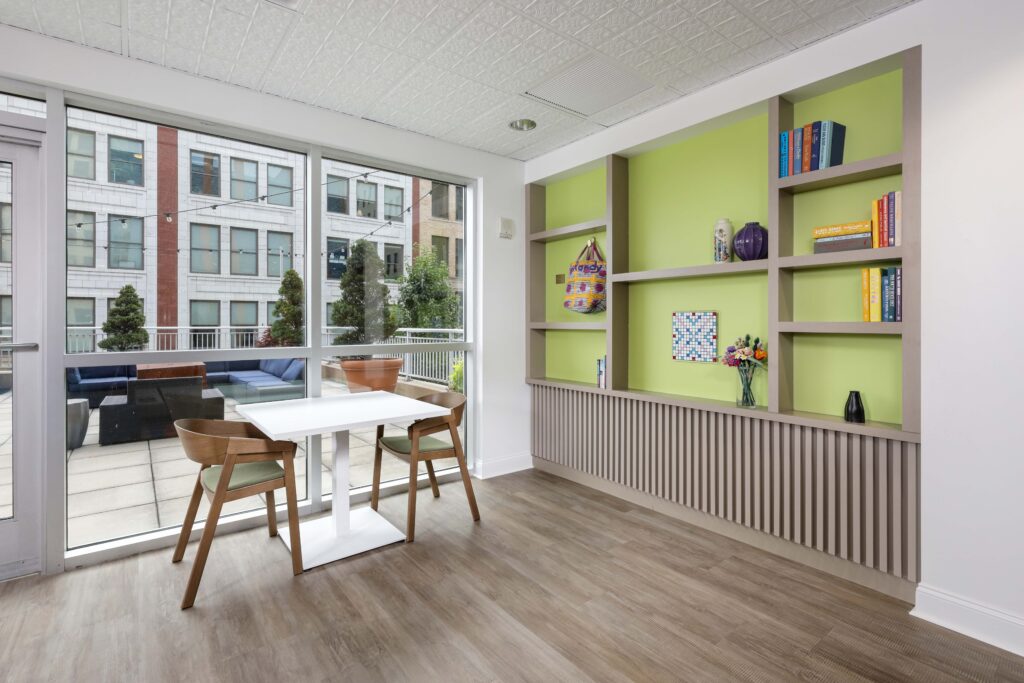 A square dining table with two chairs positioned in front of a window overlooking a patio, next to a lime-green built-in bookshelf.