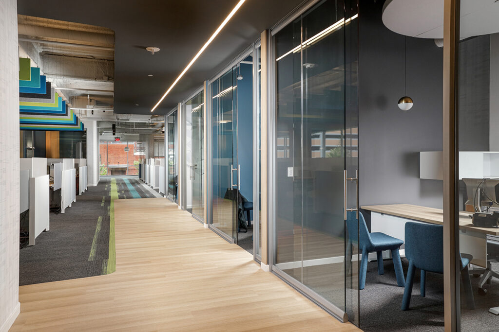 A bright office hallway featuring a row of glass-enclosed meeting rooms, a wooden floor, and open cubicle workspaces with colored acoustic panels.