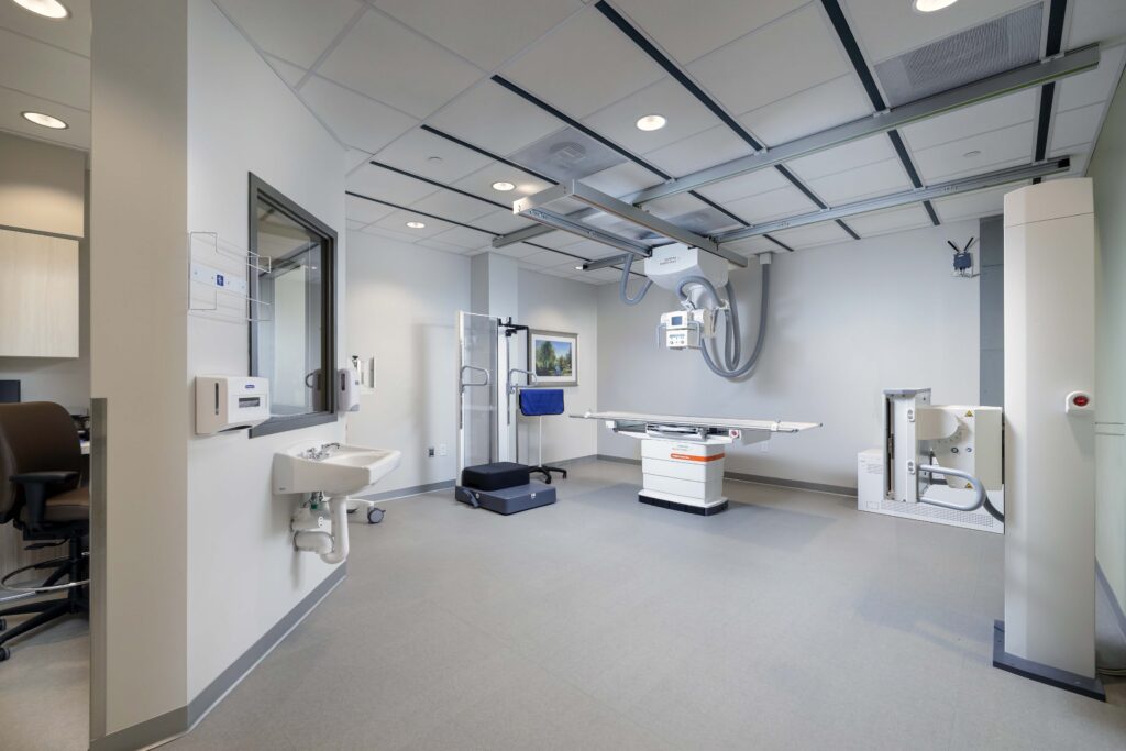 An X-ray imaging room featuring a ceiling-mounted X-ray tube over an adjustable examination table, with a standing bucky stand and a wall-mounted sink nearby.