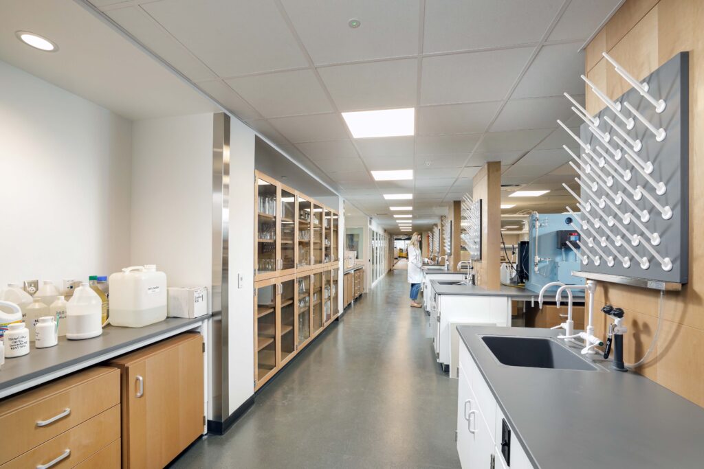 A view down a laboratory aisle with cabinets, laboratory glassware, chemical containers, and a researcher working at a bench in the distance.