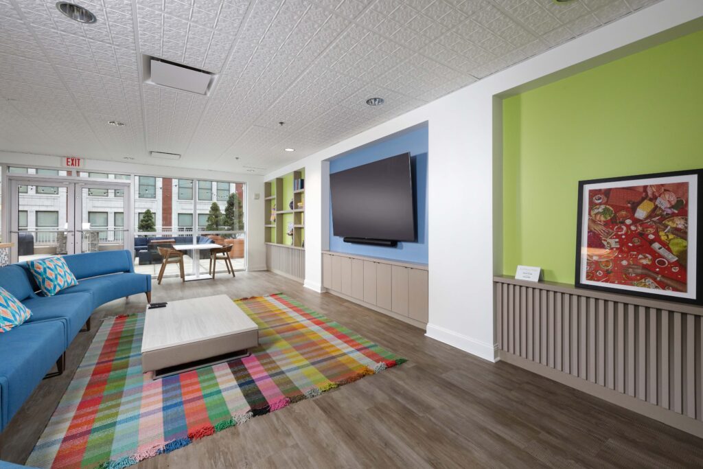 Lounge area with a blue curved sofa, coffee table, colorful plaid rug, mounted television on a blue wall, and a framed painting on a green wall.