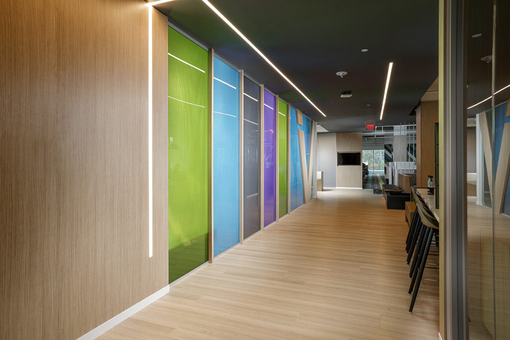 A modern office hallway with light wood flooring and walls, featuring a series of colorful translucent panels in green, blue, and purple along one side, illuminated by overhead recessed strip lighting.