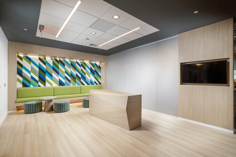 Office reception area with a wood reception desk, a green bench seat with patterned stools, and a television mounted on a wood-paneled wall.