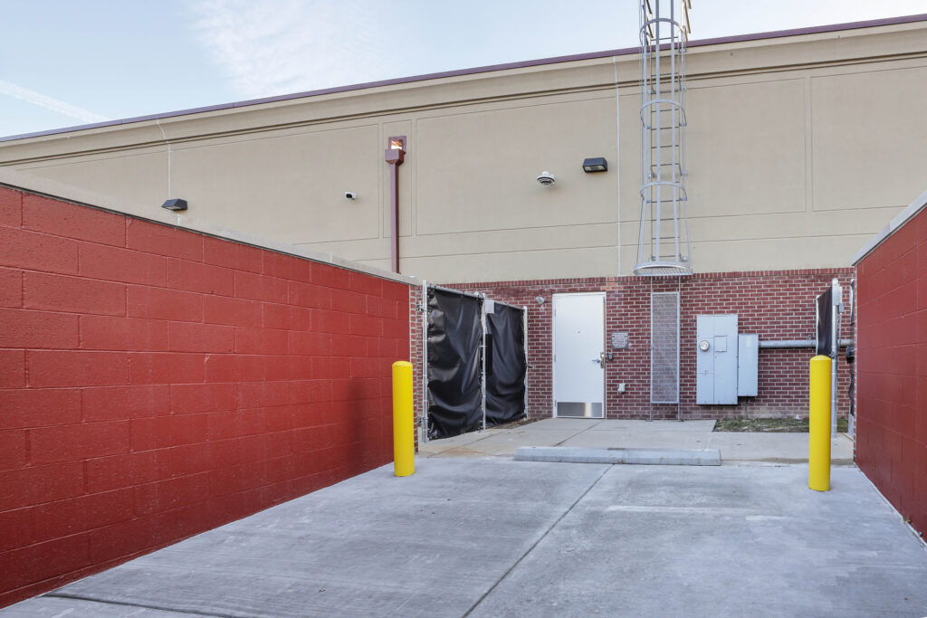 A loading bay area featuring a white door, a tall metal ladder, and two yellow bollards against red and tan walls.