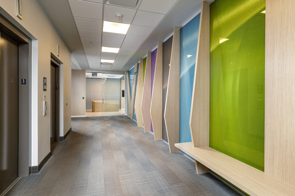 A modern office hallway with gray carpet, elevators on the left, and a long wooden bench against a wall of colorful glass panels.