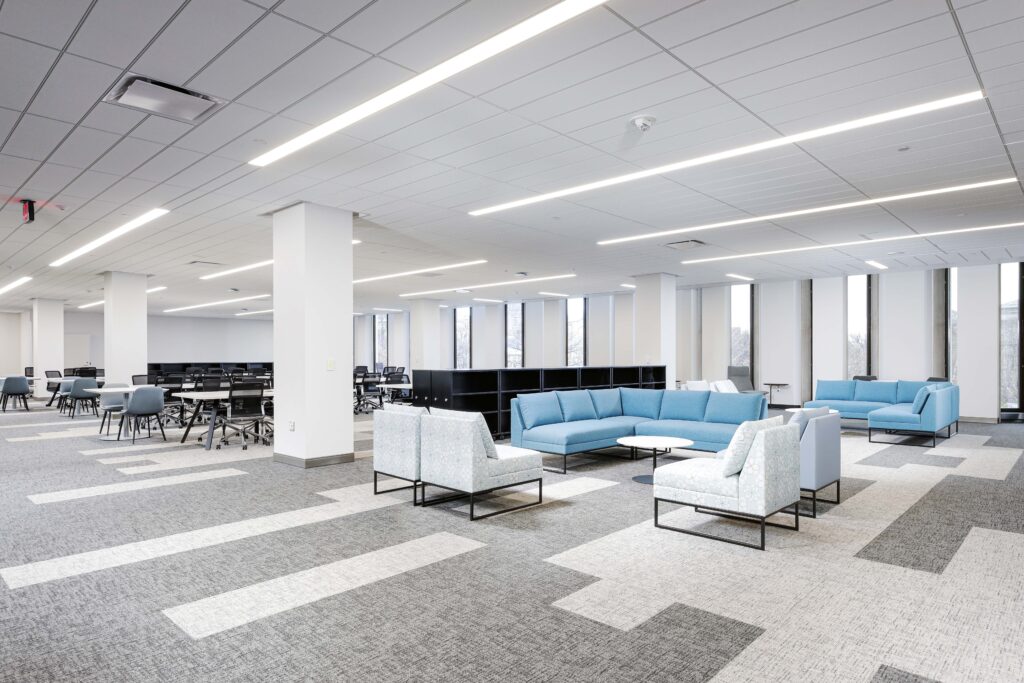 A modern open-plan office featuring a lounge area with light blue and patterned armchairs, a blue sectional sofa, and rows of desks with chairs in the background.