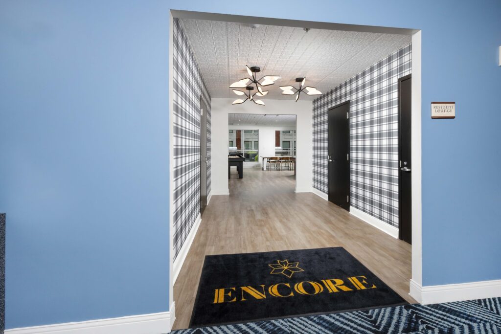 Entrance to a resident lounge featuring plaid wallpaper, a floor mat with the word “ENCORE,” and a view into a common area with tables and a pool table.