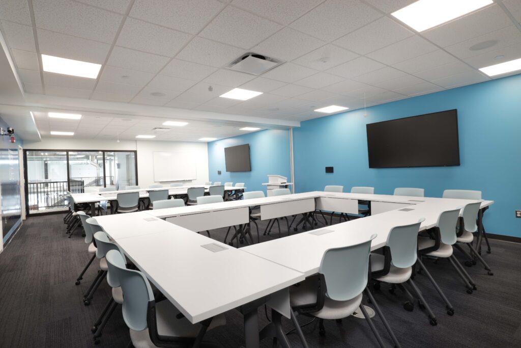 A U-shaped conference table with light blue chairs in a classroom with teal walls and wall-mounted monitors.